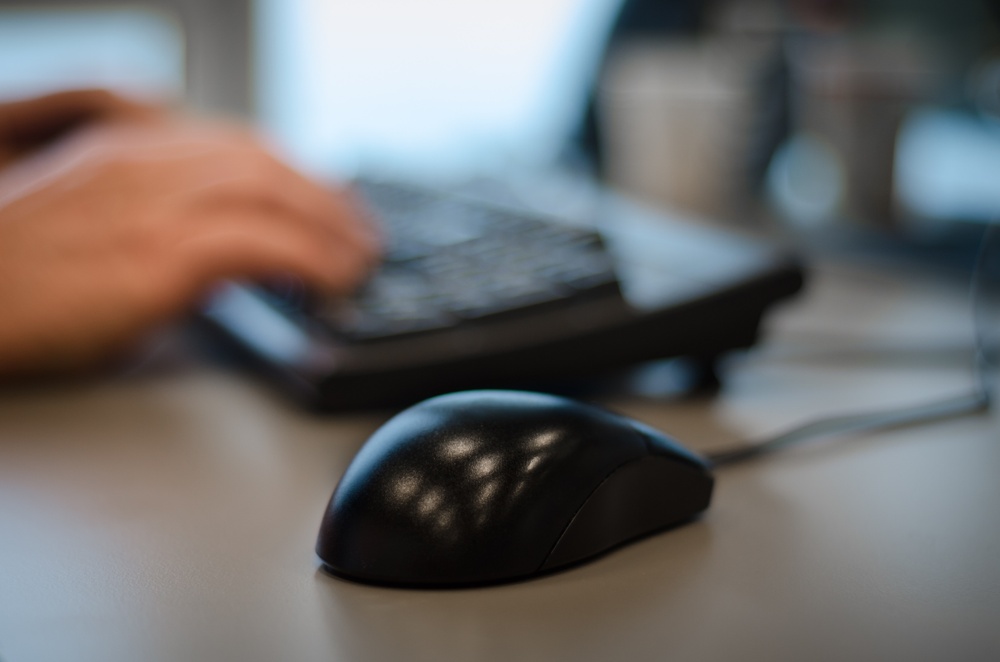 A black computer mouse in focus, and hands typing on a keyboard in the background.