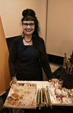 Artist Melanie Brown smiling at a work table covered in paint brushes and a paint palette