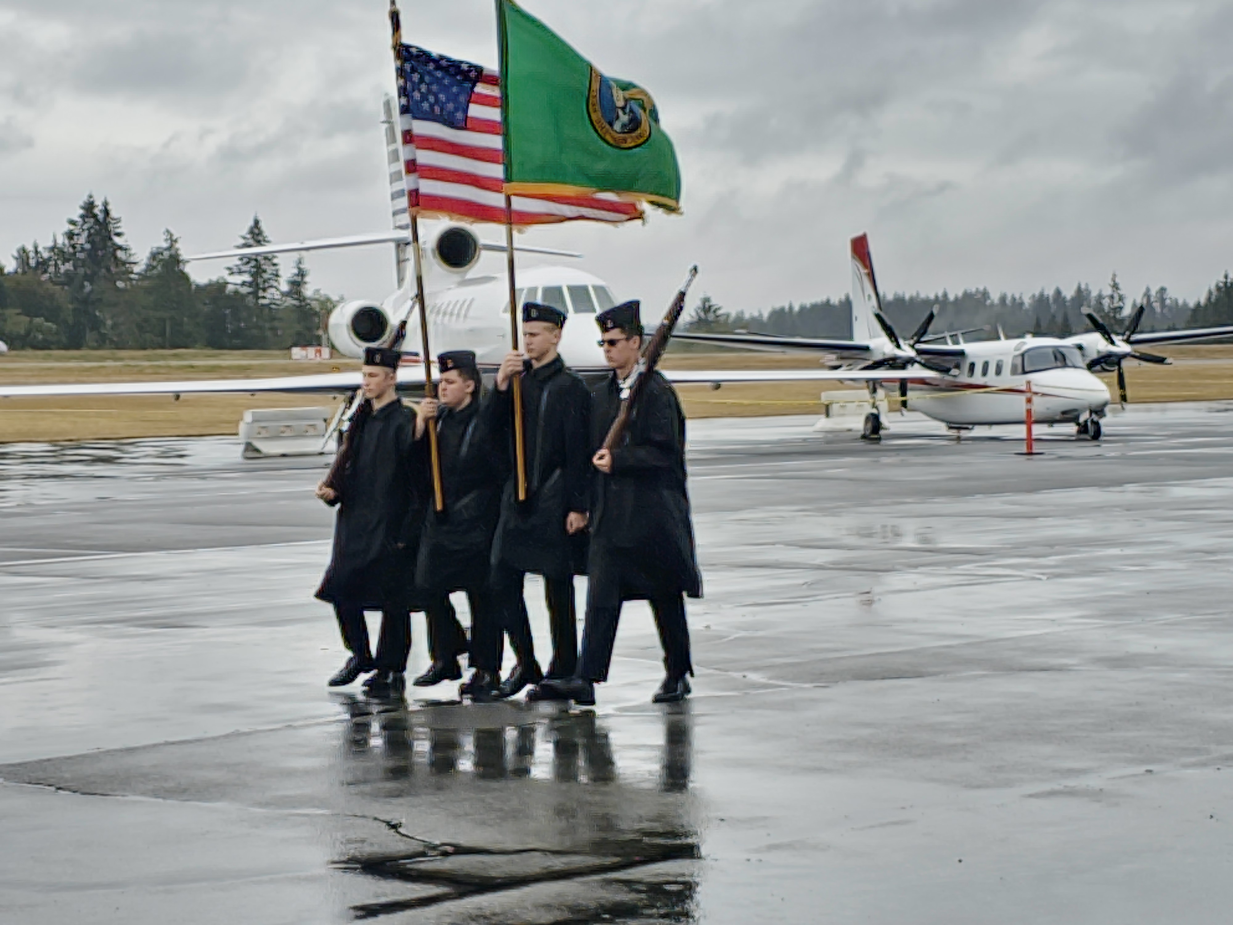 Four NJROTC students walk in step together in full uniform, with rain coats on, carrying the U.S. Flag and the Washington State flag, with two cadets carrying rifles. They are marching on a rainy day across the tarmac at a small airport ready to present the colors.