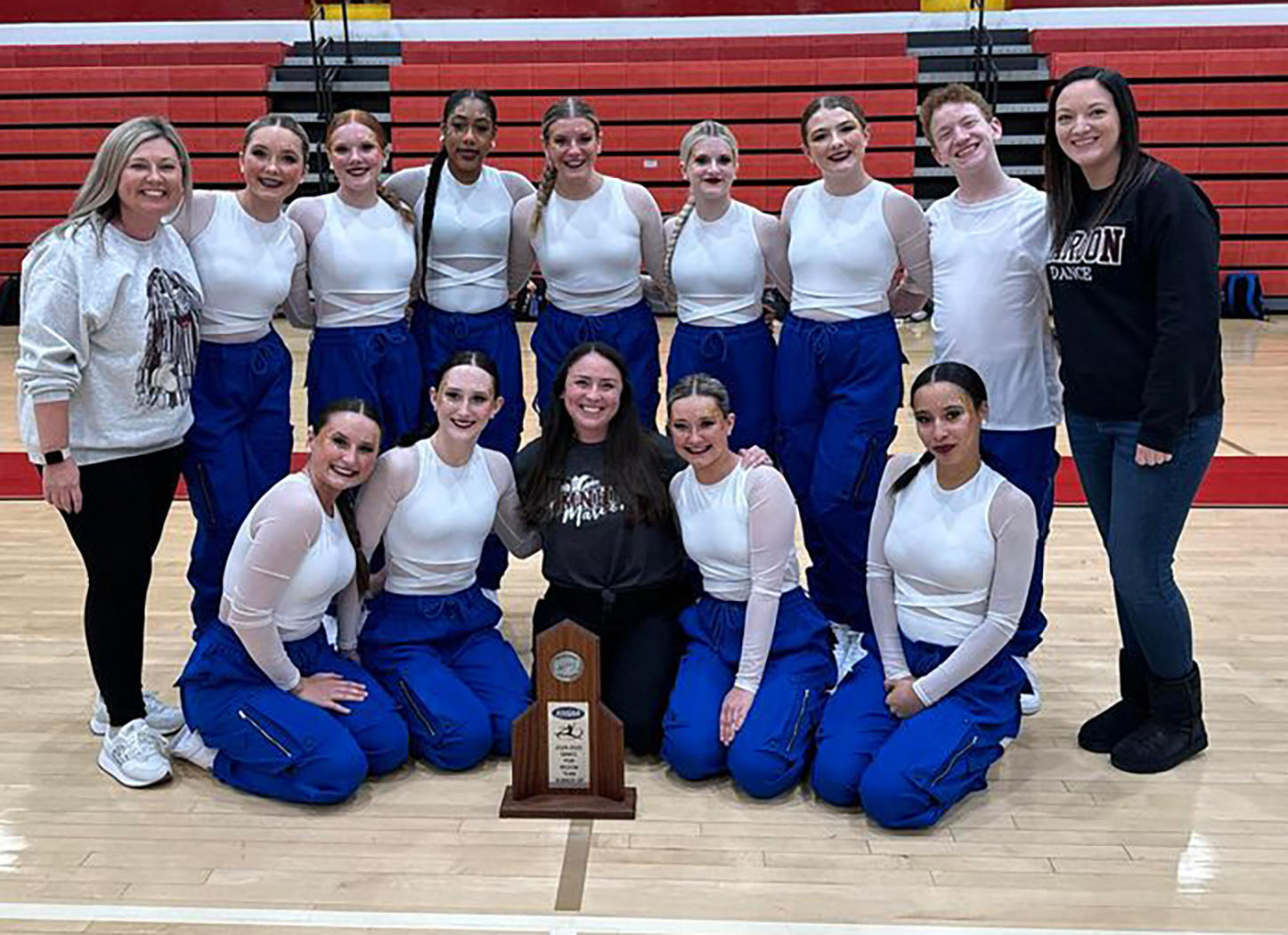 Dance team members in blue and white uniforms and 3 coaches smile for photo with trophy