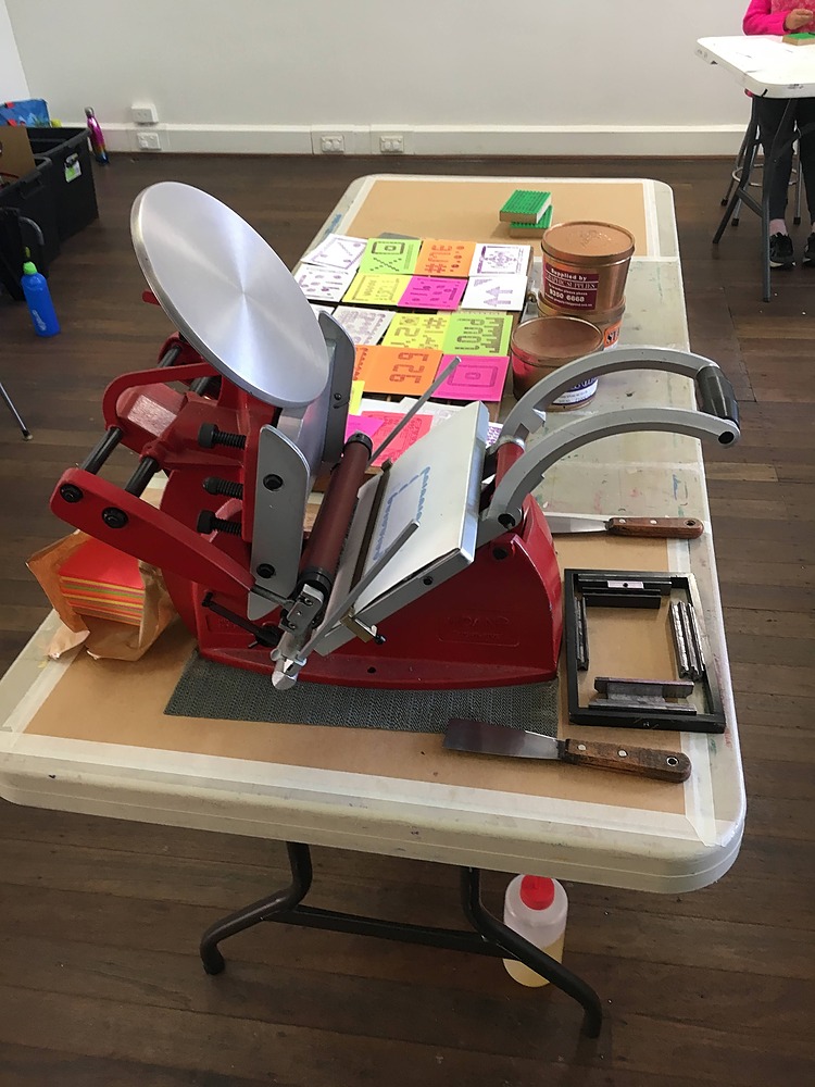 An Adana press is sitting on a picnic table. Surrounding the press are ink knives, printing chase and a stack of coloured papers. On the other side of the table are a few tins of ink and a spread of prints created by students.