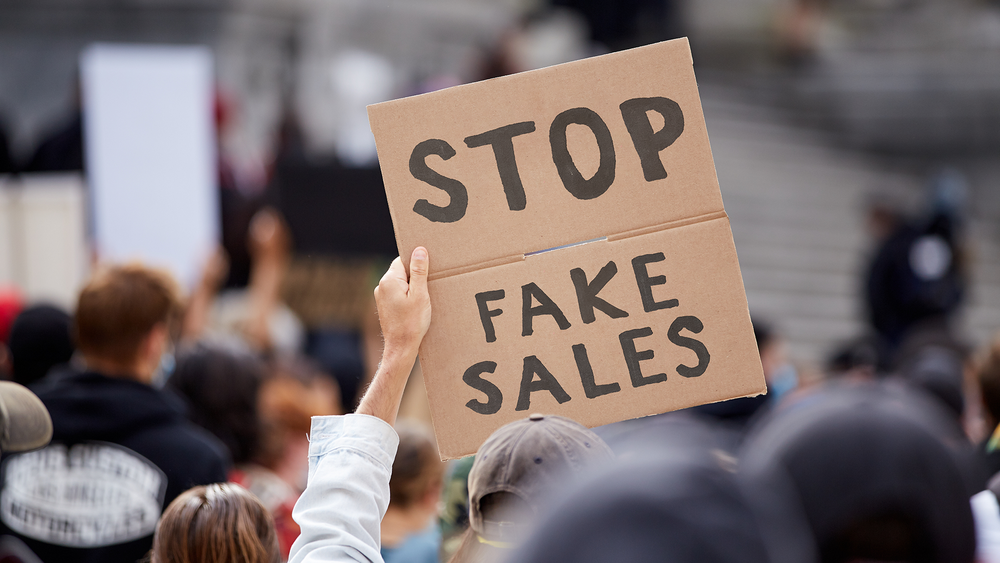 Person holding up cardboard sign which says 'Stop Fake Sales'