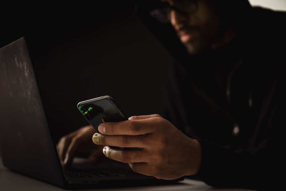 Man in a black hoodie writing on keyboard and holding a cell phone.