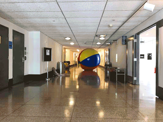 A giant beach ball inscribed with the initials "KM" rolls down the lobby of the Yale School of Art's Green Hall, while the text "News from New Haven" scrolls along the bottom in a formal newspaper-like font.