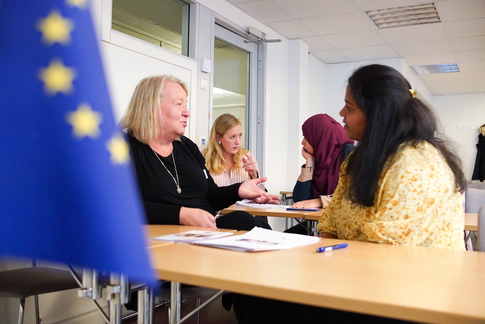 Fyra kvinnor från olika länder sitter och diskuterar med varandra i par i ett klassrum. I förgrunden syns EU-flaggan.