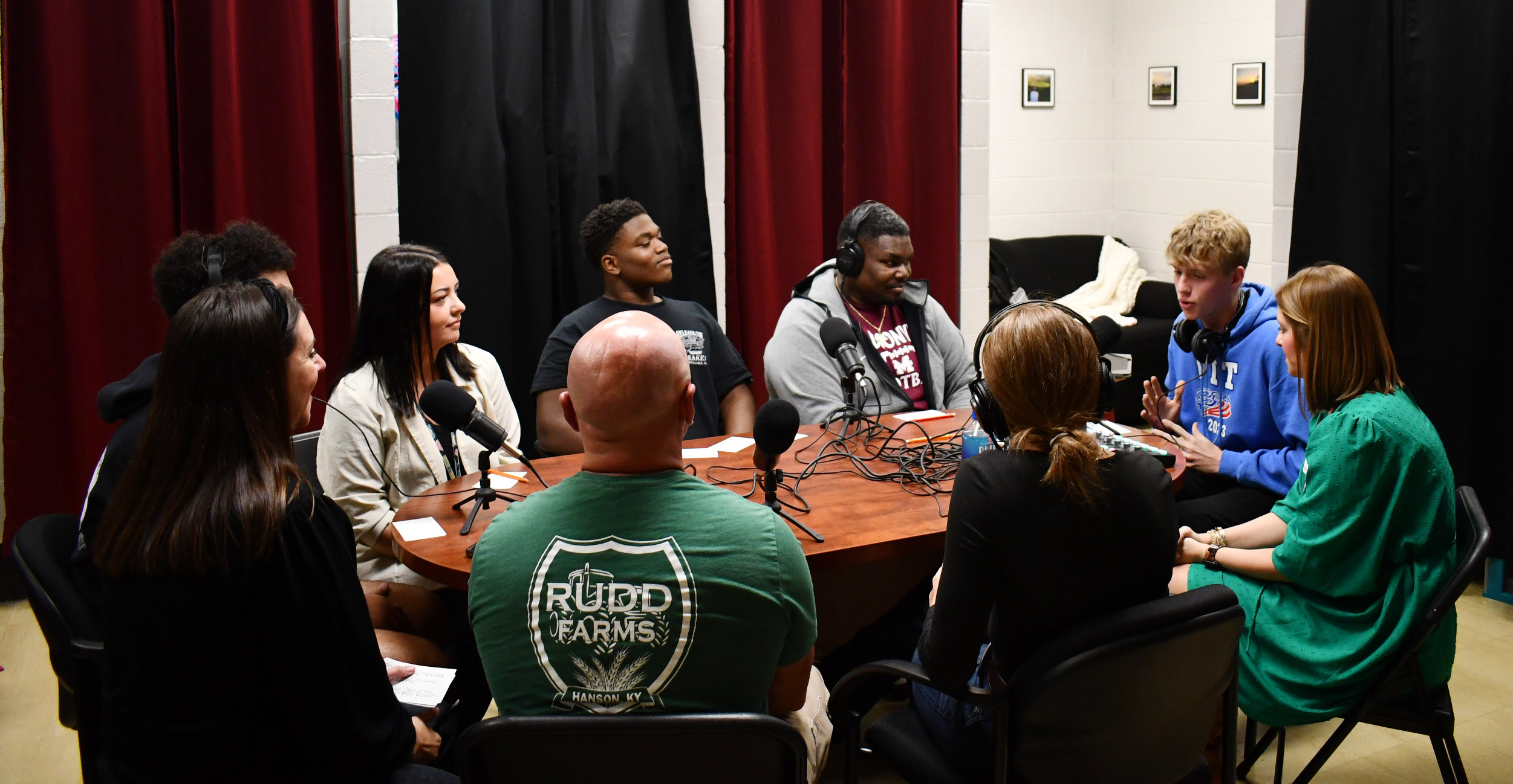 9 people sit at table with recording equipment on it.