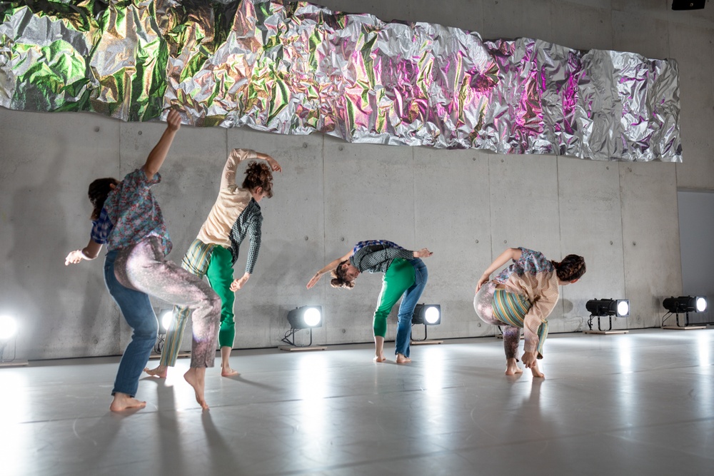 Fyra dansare rör sig på en scen med ljust grå betongväggar och golv. I fonden hänger en foliefärgad duk med påmålade pastellfärger i gröna och rosa toner.
