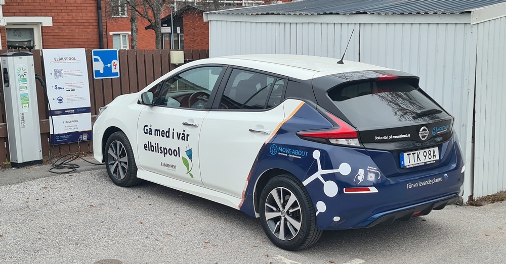 Den vita bilen med fyra dörrar och blåfärgad baktill står parkerad och laddas med förnybar energi intill ett bostadsområde i Upplands Väsby.