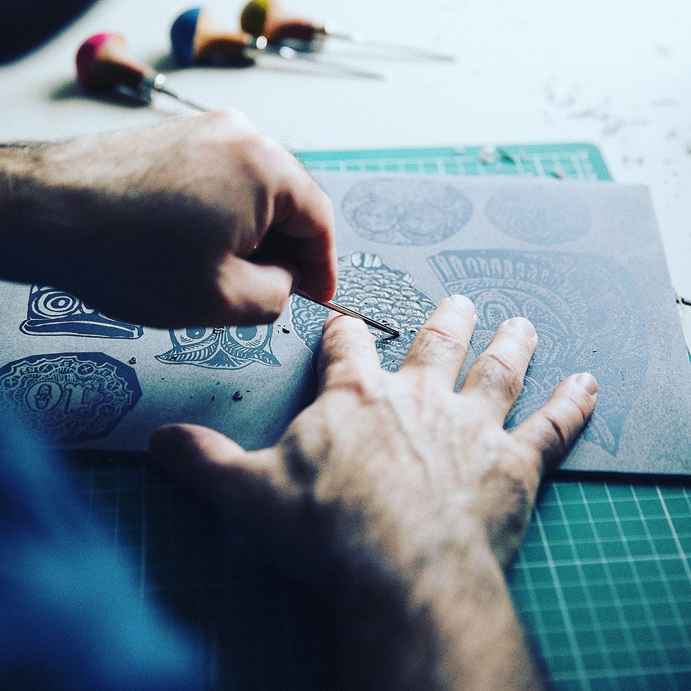 A pair of hands is carving into a lino block with a carving tool on the left hand and the right hand is supporting the block.