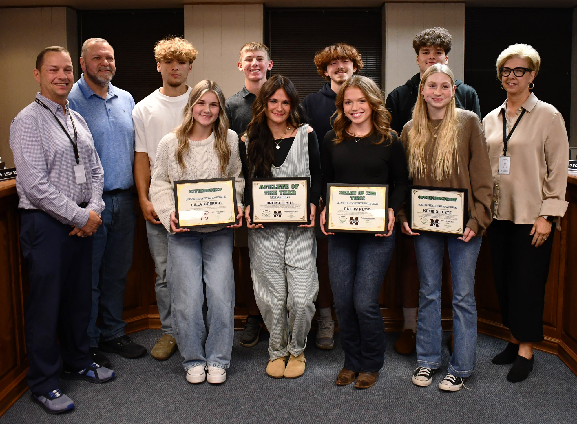 11 people stand and smile for photo, with 4 of them holding framed certificates.