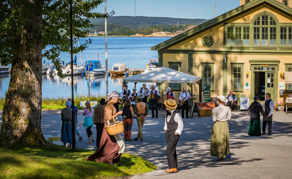 Bild. Framför det gula varmbadhuset på Gustafsberg vandrar ett flertal människor iklädda tidstypiska kläder från 1800-talet.