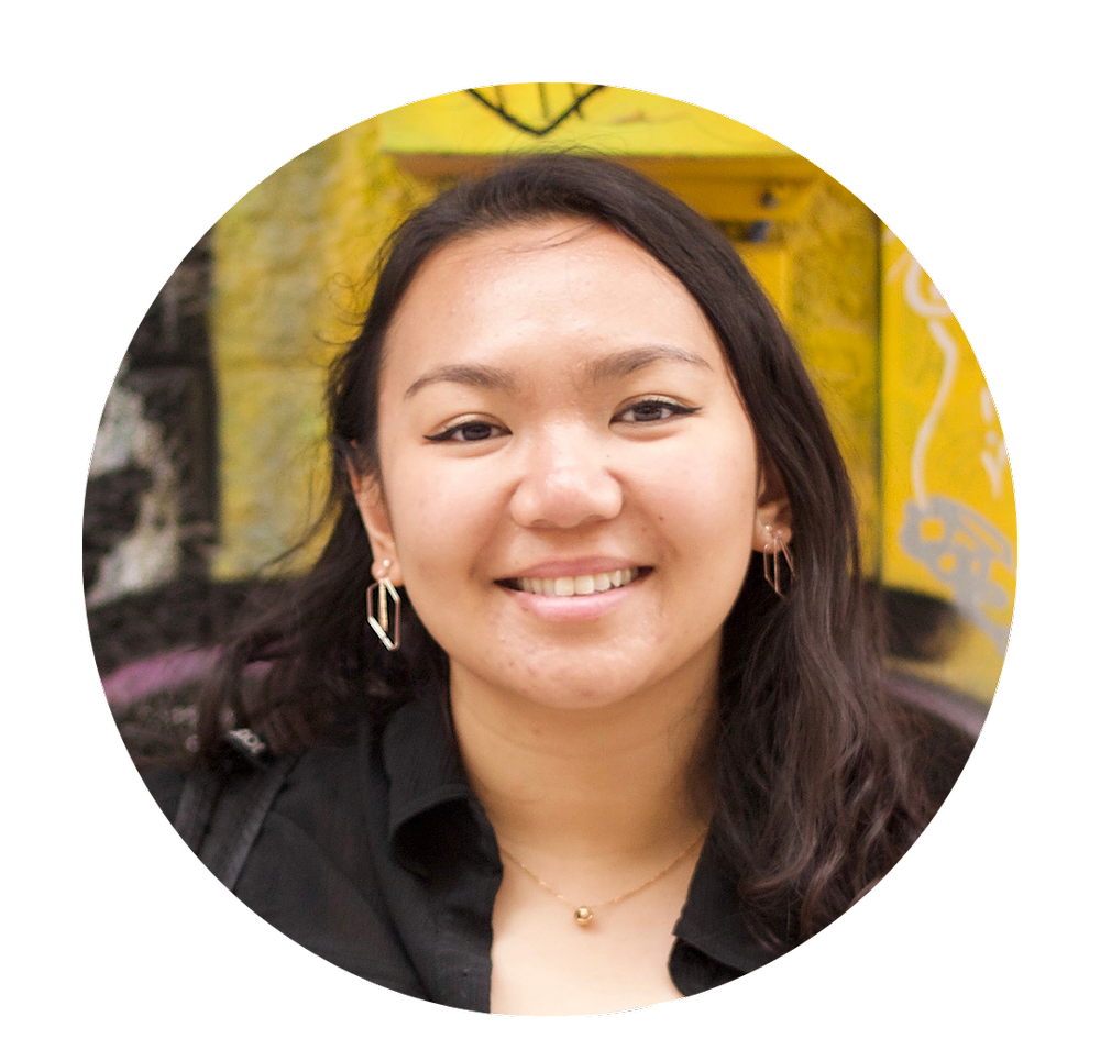 A Southeast Asian woman with black hair in a black top is smiling with teeth showing, with a yellow wall in the background.