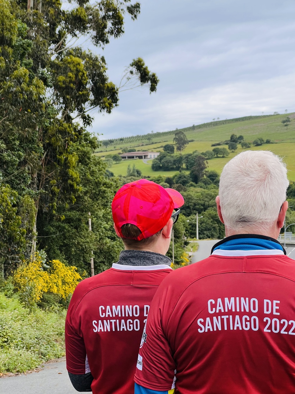 Två personer med ryggen mot kameran, de har röda tröjor där det står Camino De Santiago 2022 på ryggen.