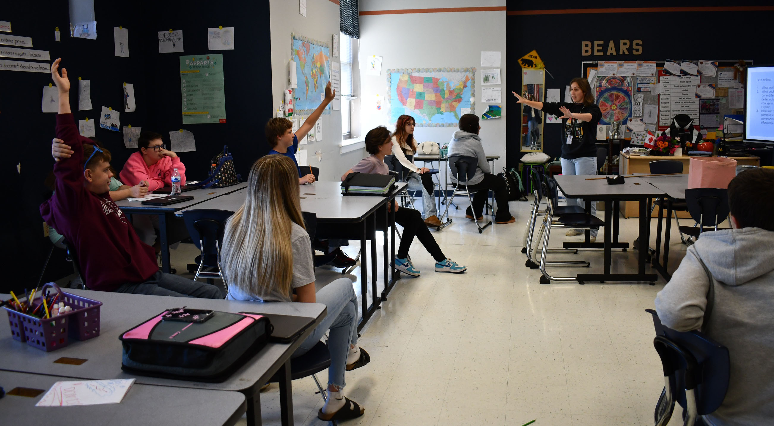 Teacher stands at front of classroom with about 8 students seated at desks and the word "bears" on the wall.