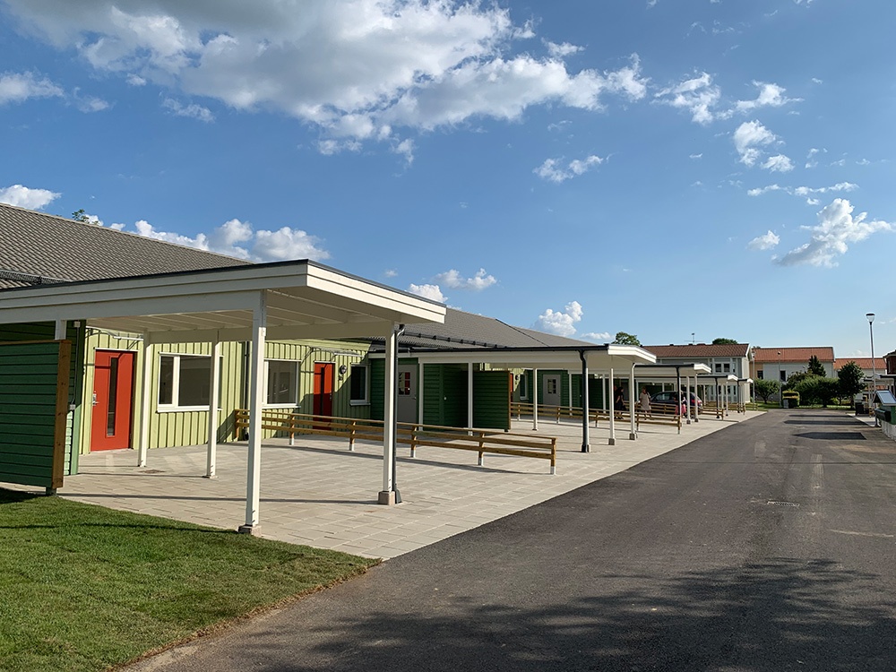 Grönt hus utmed gata. Framför vare röd lägenhetsdörr finns en stor uppfart med uteplats och carport.