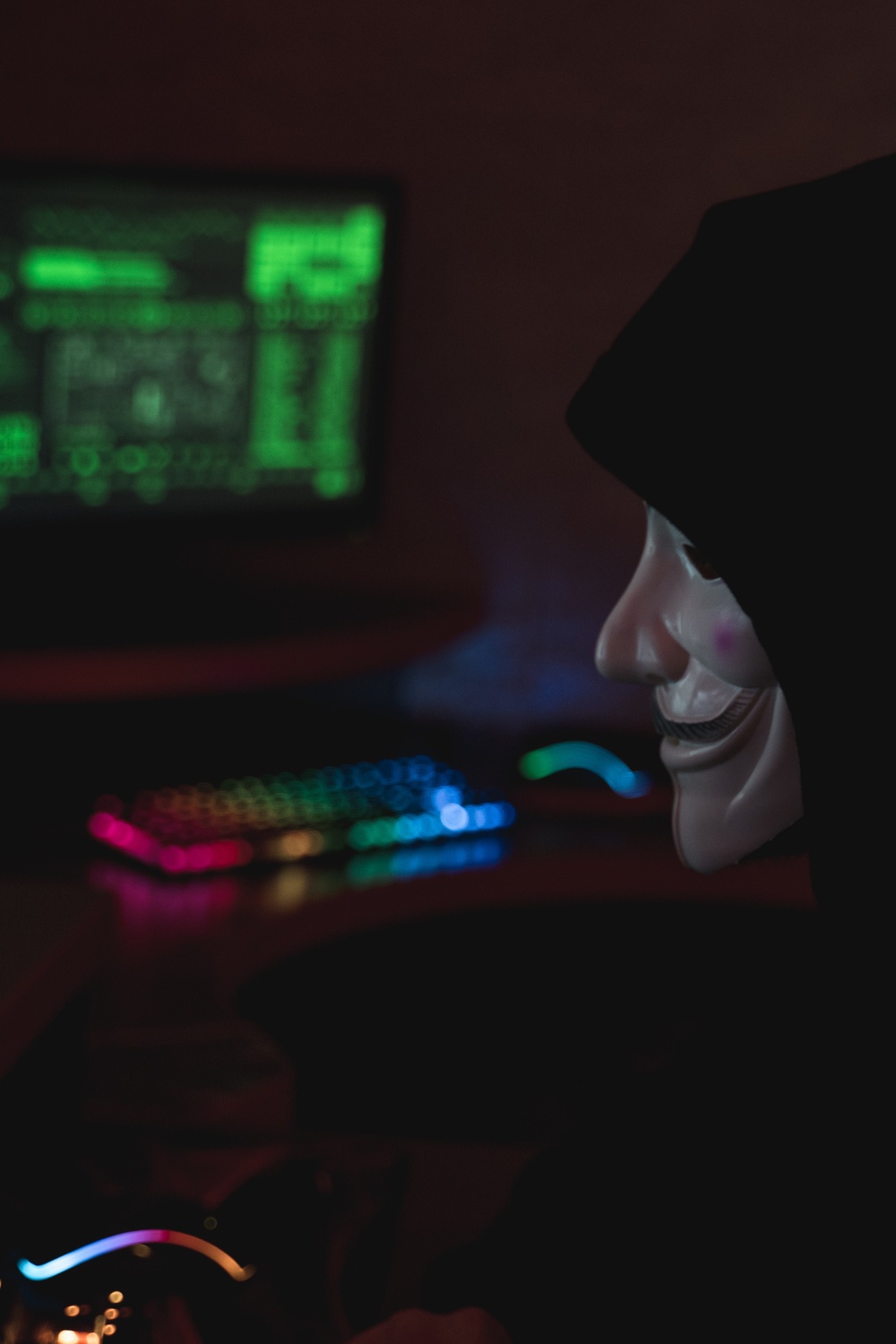 A dark room with a computer screen, green letters on the screen and a hooded figure wearing an Anonymous/Guy Fawkes mask.