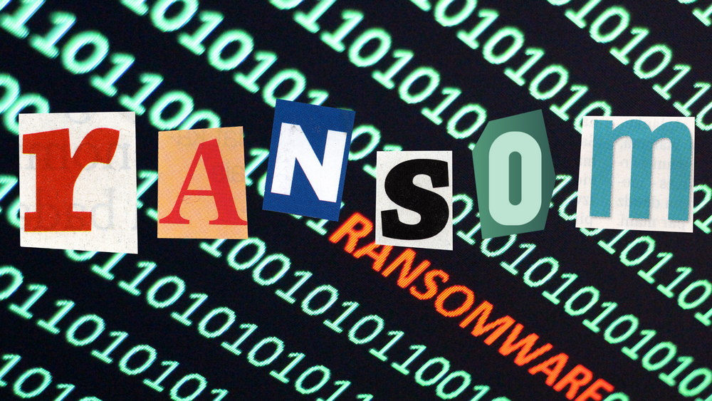 Green ones and zeros on a computer with the text "RANSOMEWARE" highlighted in orange. The letter "RANSOM" are cut out from a nesapaper and sits in front. 