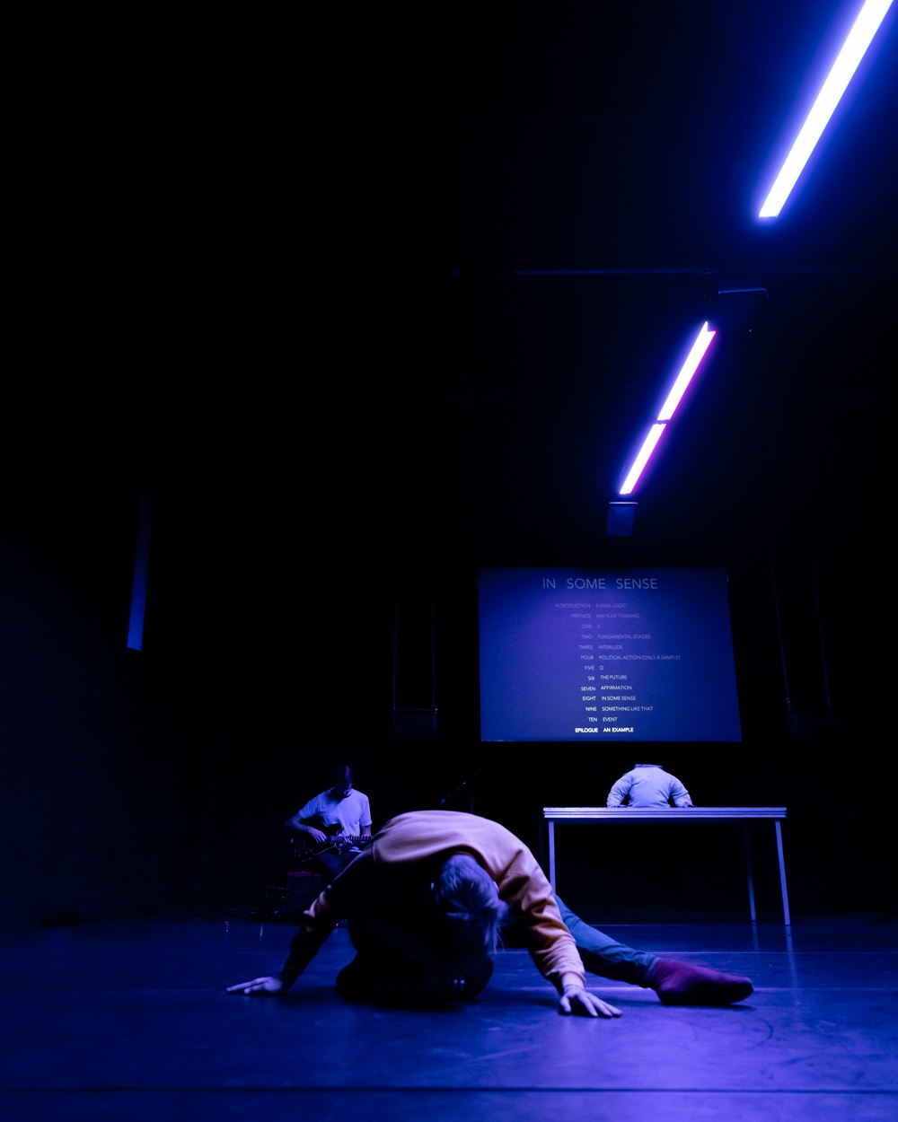 En mörk scen med svagt blått ljus som strilar ner. I bakgrunden en projektionsduk med text. Längst fram ligger en manlig dansare. I bakgrunden skymtar en gitarrist.