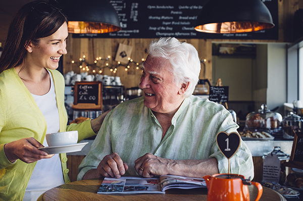 A very happy older man is sitting at a table in a cafe with a magazine open in front of him. He is smiling at the woman bringing him his hot drink.