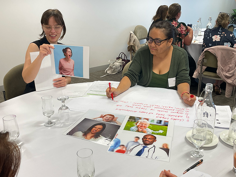 2 women are seated at a table. One is holding up a photo of a person and the other is writing on butcher's paper. There are other photos of people representing different professions on the table