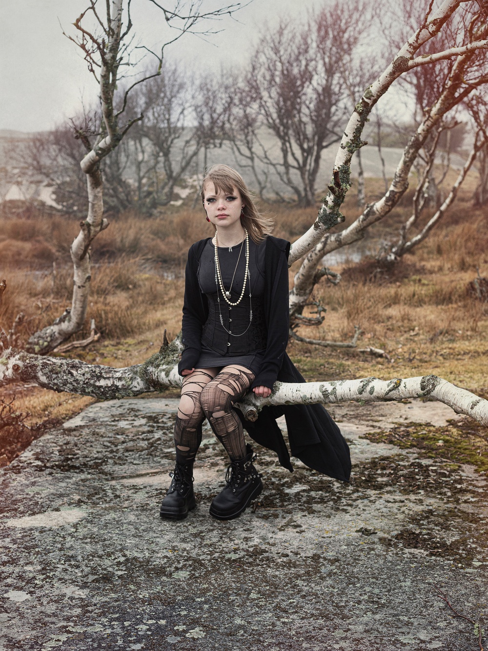 Foto. I ett höstigt kustlandskap sitter en ung blond kvinna på en björk. Hon bär svarta kläder, trasiga svarta strumpbyxor, svarta boots och flera långa halsband runt halsen. 