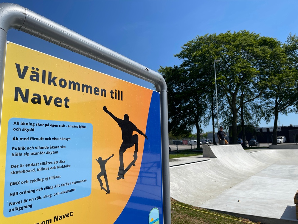 Välkomstskylt i orange och blått med ordningsföreskrifter. Skateboardbanor och träd i bakgrunden