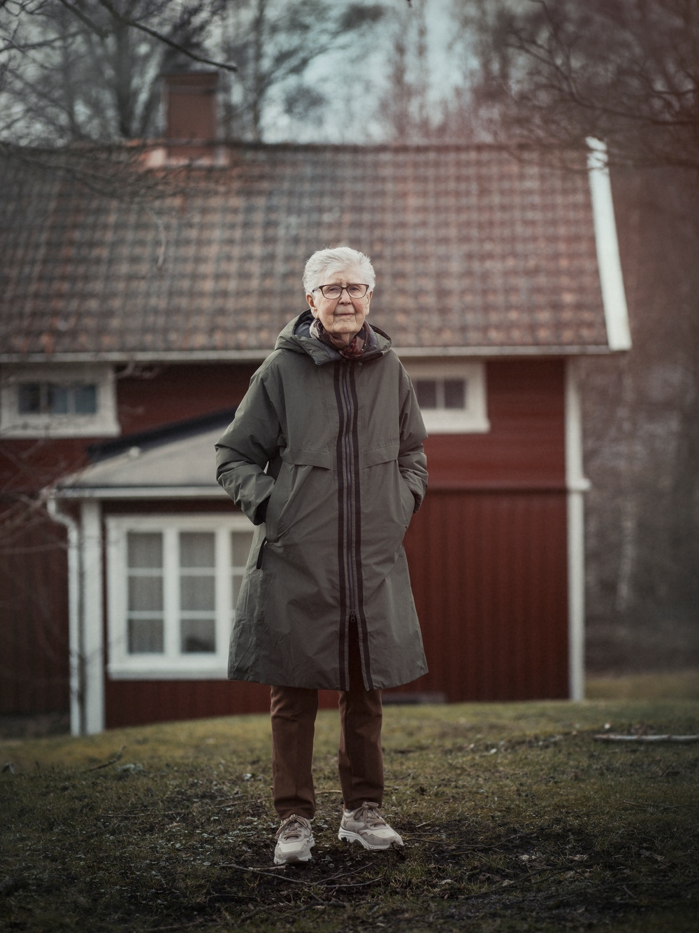 Foto. En vithårig, kortklippt kvinna står i sin trädgård iklädd en lång täckjacka. I bakgrunden syns ett rött torp.