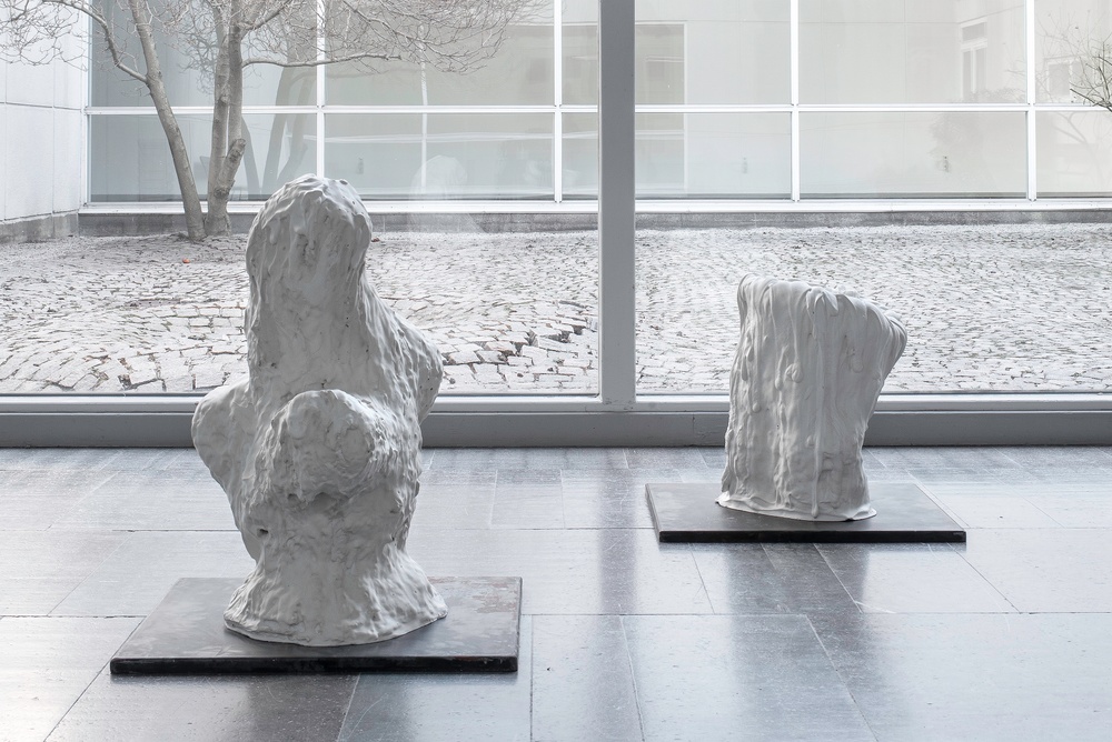 Two stump-like white sculptures on low plints stand on a stone floow. In the backround large windows opening to a cobbled atrium of a Modernist building.