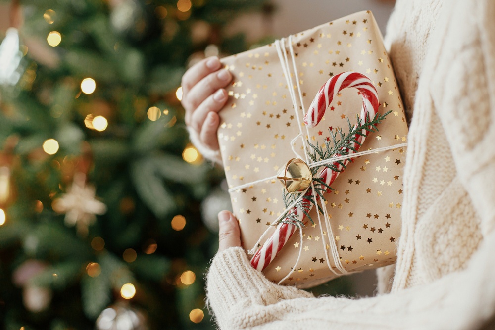 Woman with a white sweater holding a gift with a peppermint cane on it. There is a Christmas tree in the background