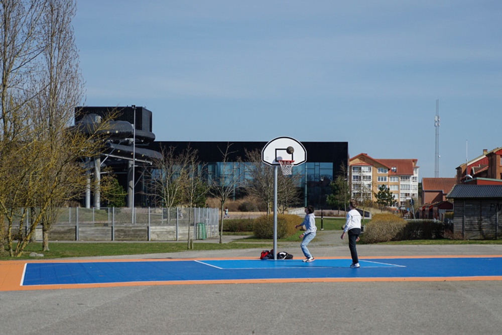 Två personer spelar basket i Åvallaparken, med Ystad Arena Bad i bakgrunden