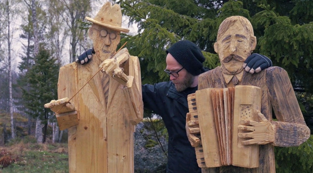 En man står utomhus mellan två träskulpturer.