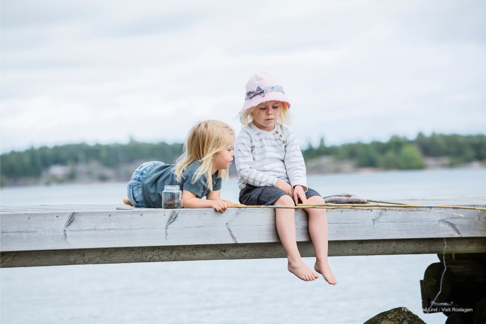 Två barn sittandes på en brygga med fiskespön i hand och en maskburk intill. 