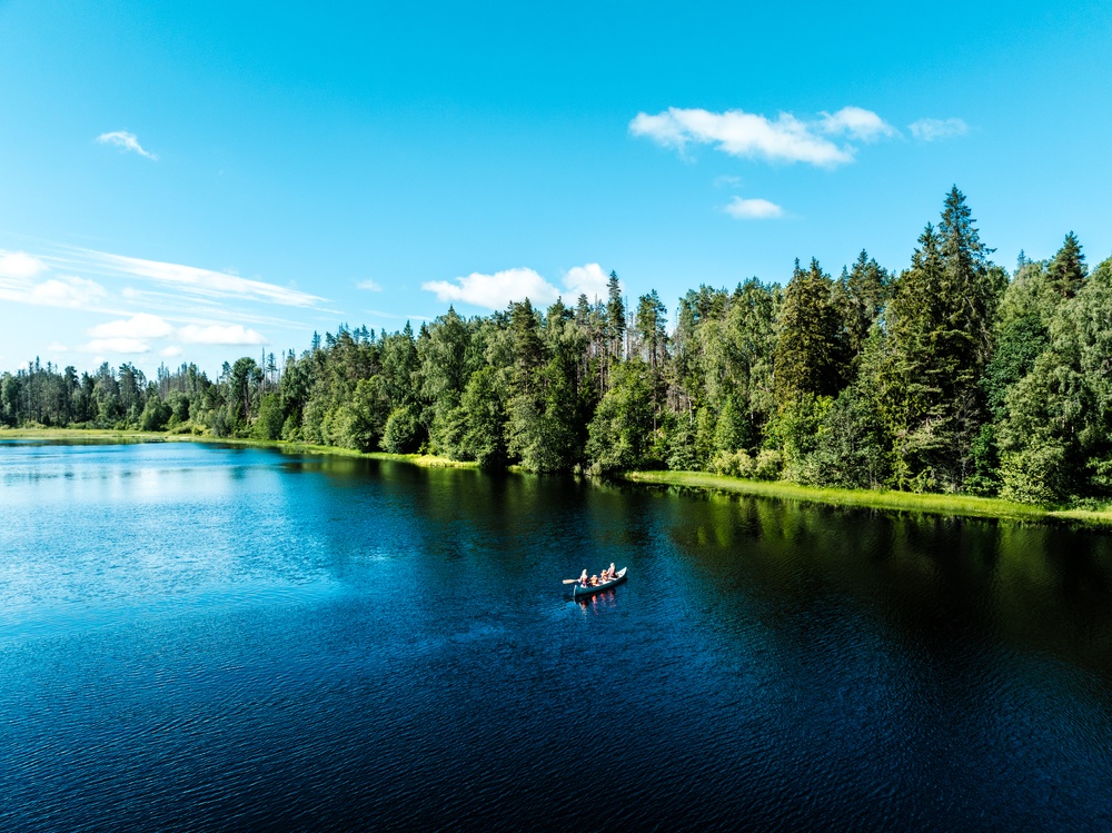 Drönarbild över en stilla sjö med grön barrskog vid stranden och blå himmel. En kanot på sjön. 