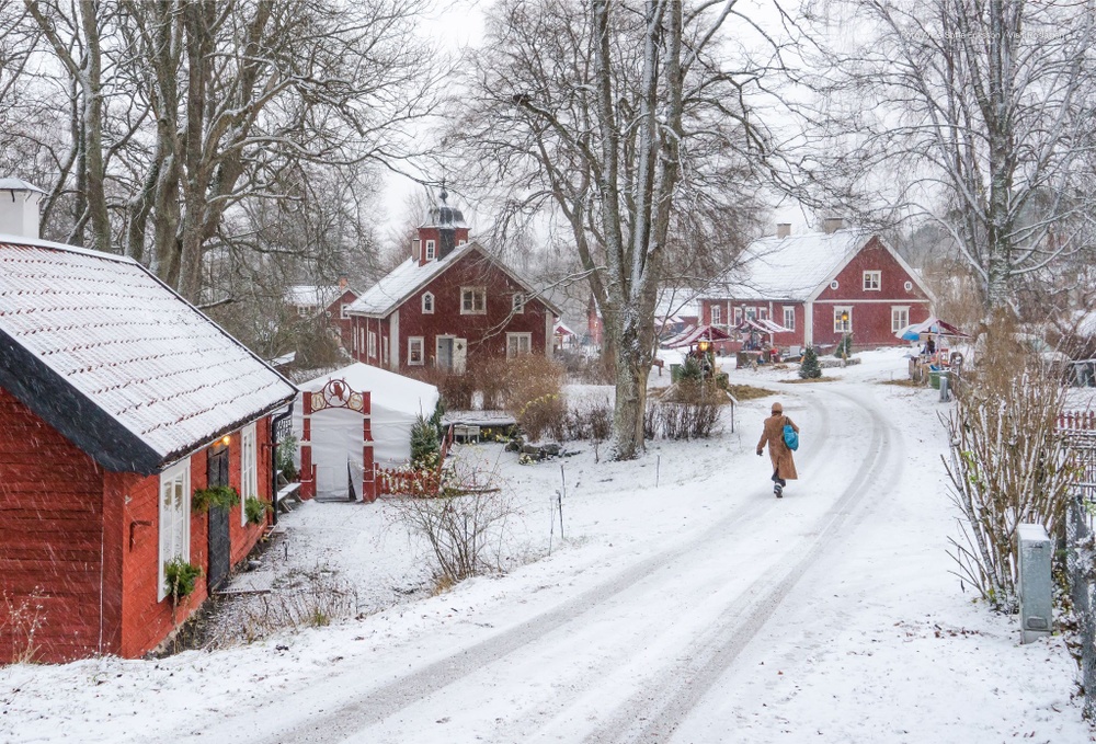 Röda stugor, snötäckt väg och en kvinna gåendes mot julmarknadsknallar.