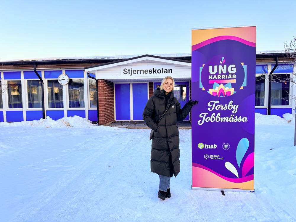 Vy från utsidan Stjerneskolan med skolbyggnaden i bakgrunden. En färgglad rollup med Ung Karriär - Torsby Jobbmässas logotype. Elvira Berglund, projektledare, poserar till vänster om rollupen. 