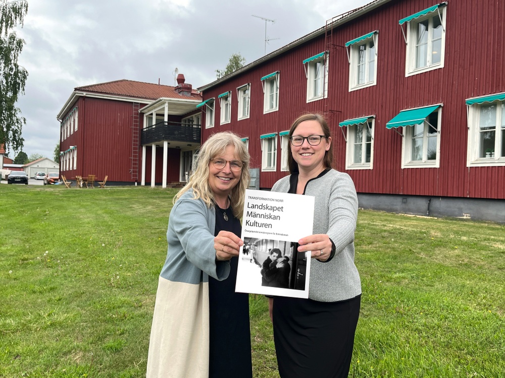 Två kvinnor håller tillsammans fram en broschyr. I bakgrunden ett rött stort hus.