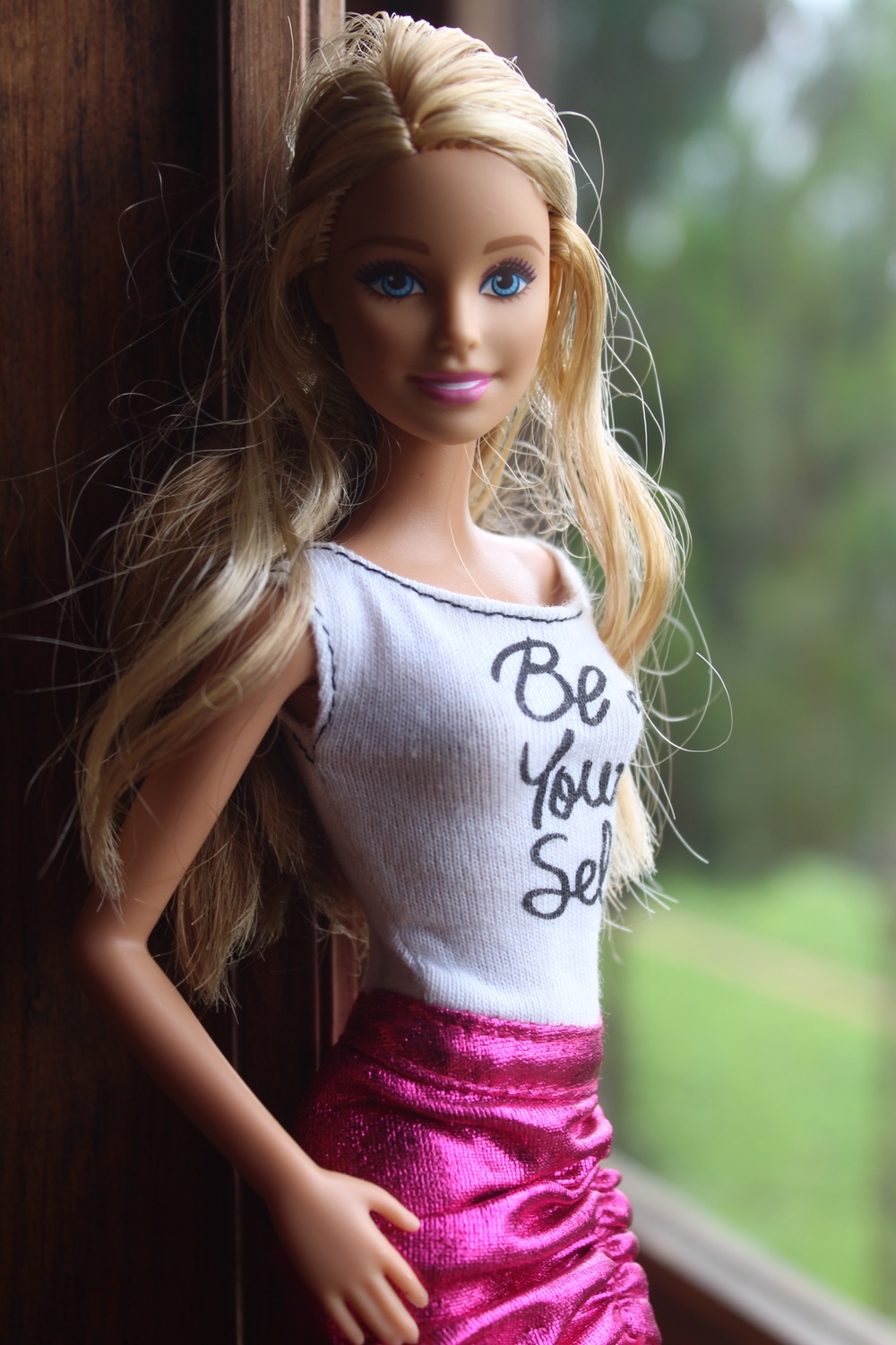 Vaaleahiuksinen Barbie seisoo ikkunan edessä valkoisessa paidassa ja pinkissä hameessa. 