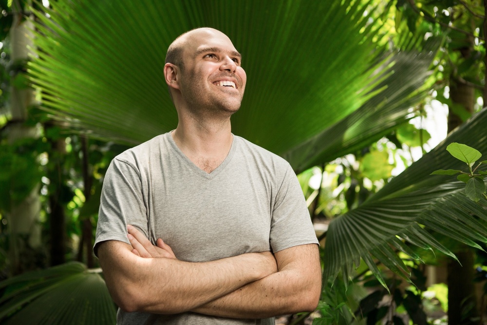 Artist David Elia stands in front of lush, tropical plants. The artist draws inspiration from nature and landscape.