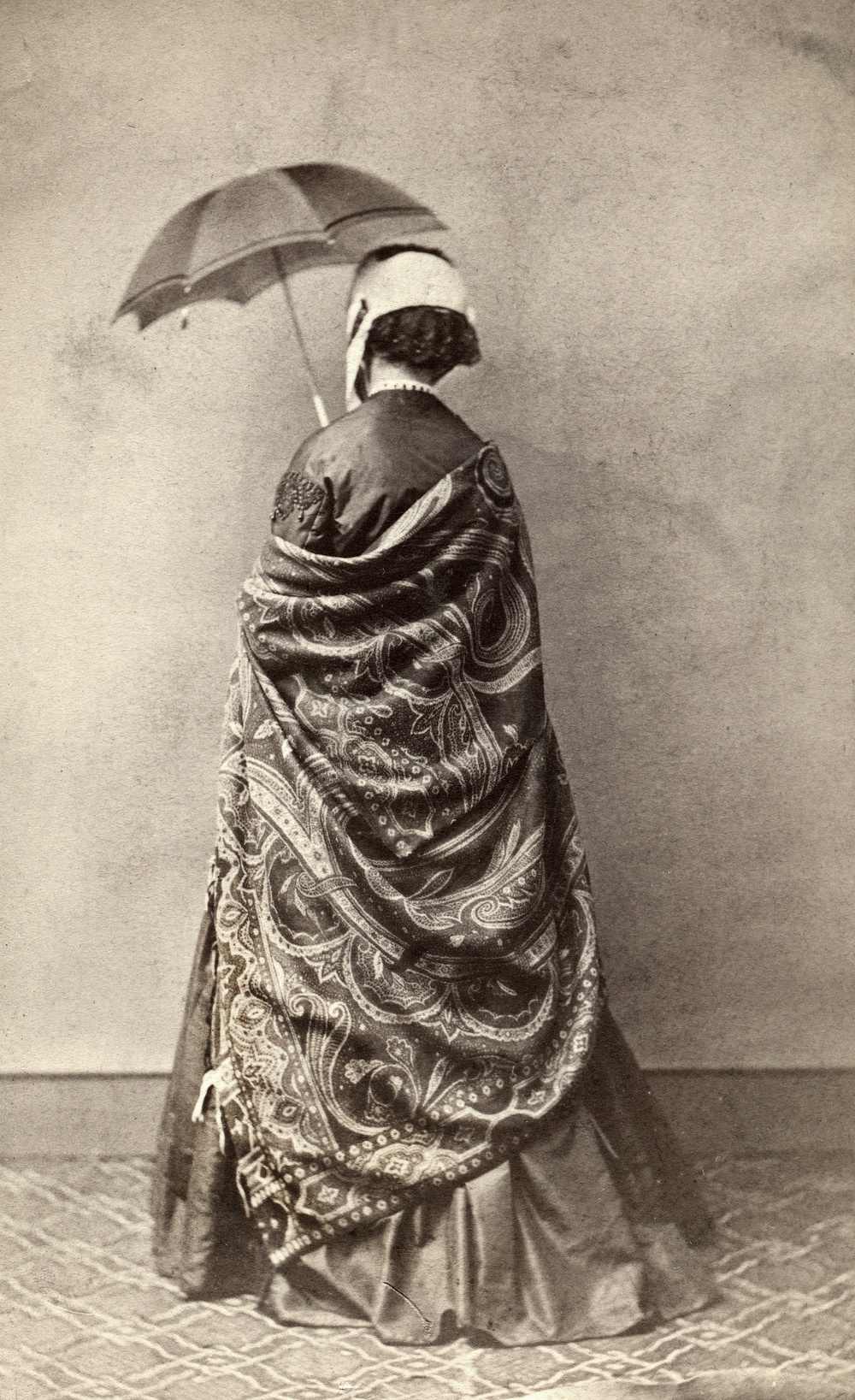 Fotografi från 1860-talet med en kvinna i stor, mönstrad schal, mörk sidenklänning, ljus hätta med svart spets och parasoll. Enligt texten på baksidan vägrade hon att visa sitt ansikte: ”alla böner och öfvertalningsförsök otillräckliga”.                                                                                                                 Fotograf Hilda Thysenius, Ronneby, 1860-talet. Gåva av Annie Olins sterbhus 1961. 
Albuminsilverfotografi monterat på kartong. Visitkort.
