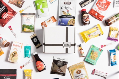 A subscription box labeled The Keto Box near keto-friendly snacks including chocolate syrup, pork panko, and flax crackers.