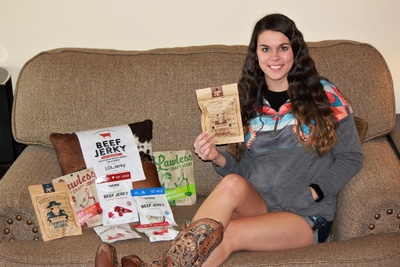 A woman sitting on a couch holding a pack of beef jerky, next to a bunch of products from a LOLJerky subscription box.