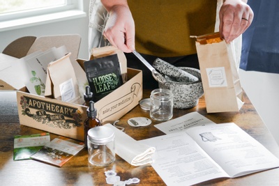 An Herbal Apprentice subscription box sitting on a table, filled with loose leaf tea packets. A person is filling packets.
