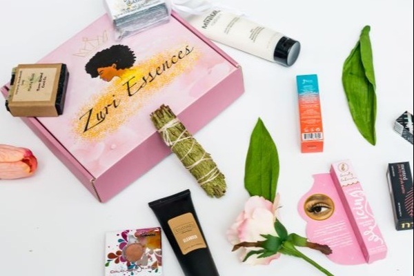 A Zuri Essences subscription box surrounded by products including a sage bundle and deluxe makeup and skincare products.