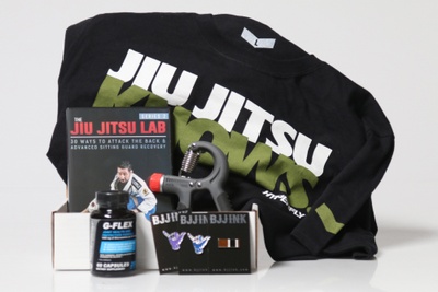 An open BJJ subscription box filled with a t shirt, a Jiu Jitsu Lab DVD, a hand strengthener, and G-Flex supplements.