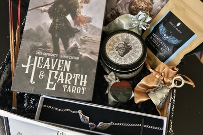 An open Nine of Earth subscription box filled with a heart and wing necklace, tarot cards and a meditation and spell kit.