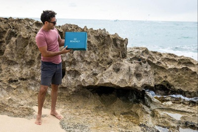 A man standing on a rocky beach, holding a closed Big Kahuna seasonal subscription box.