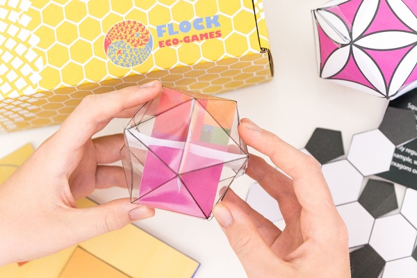 Hands holding an origami hexagon shape and a yellow Flock Eco Games subscription box.