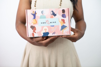 A close-up photo capturing a woman's hands cradling a HER-MINE box, which is adorned with various images of diverse women.