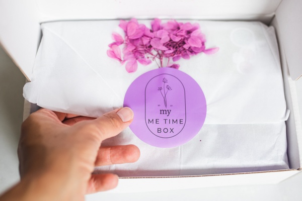 An open My Me Time subscription box with a hand opening the tissue paper with a purple flower and label on it.