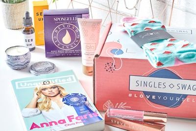 A closed SinglesSwag subscription box with socks on top of it and makeup, skincare items and a magazine next to it.